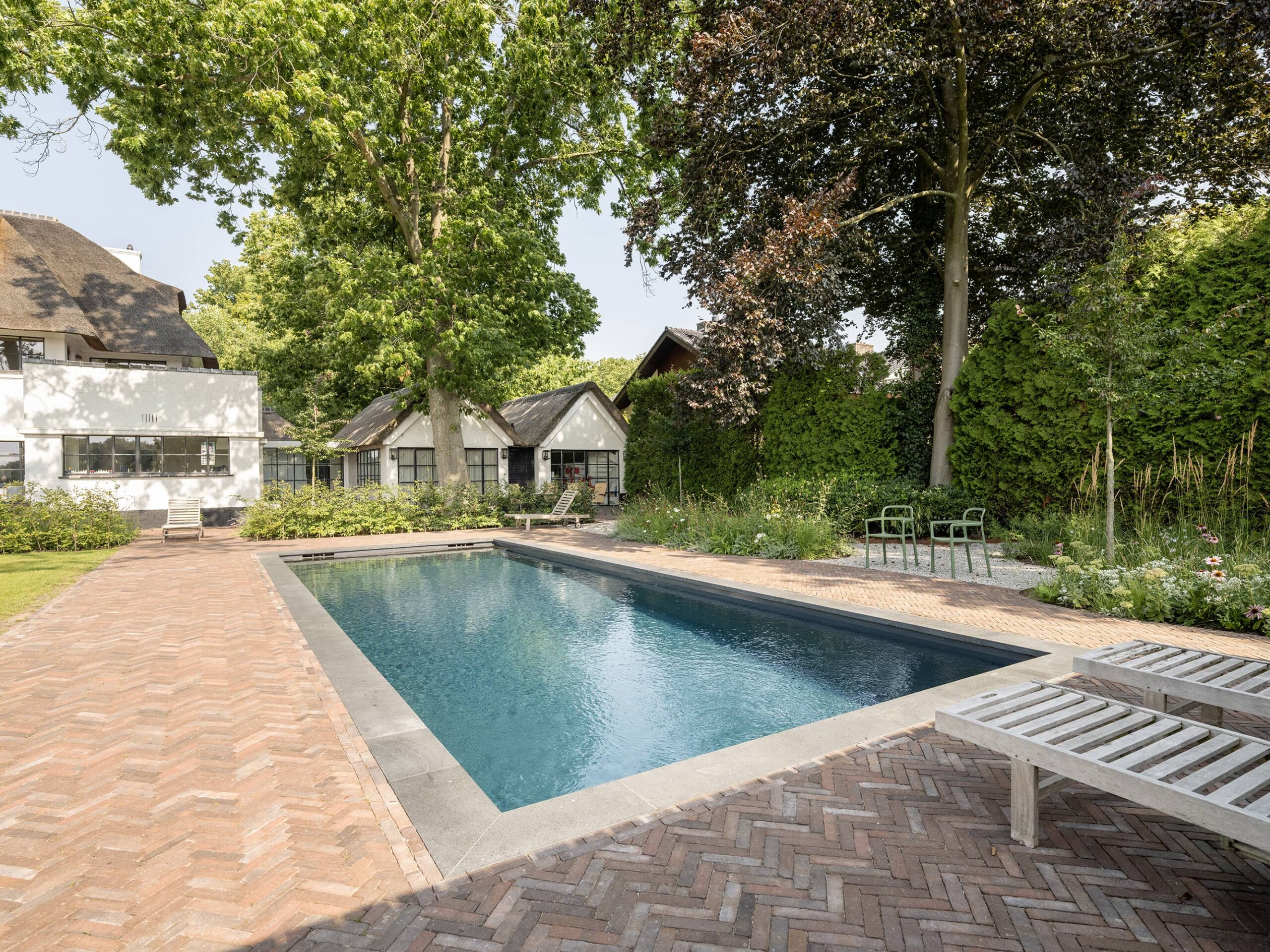 Modern buitenzwembad in ruime groene tuin met visgraat bestrating, ligbedden, volwassen bomen en witte villa op de achtergrond.